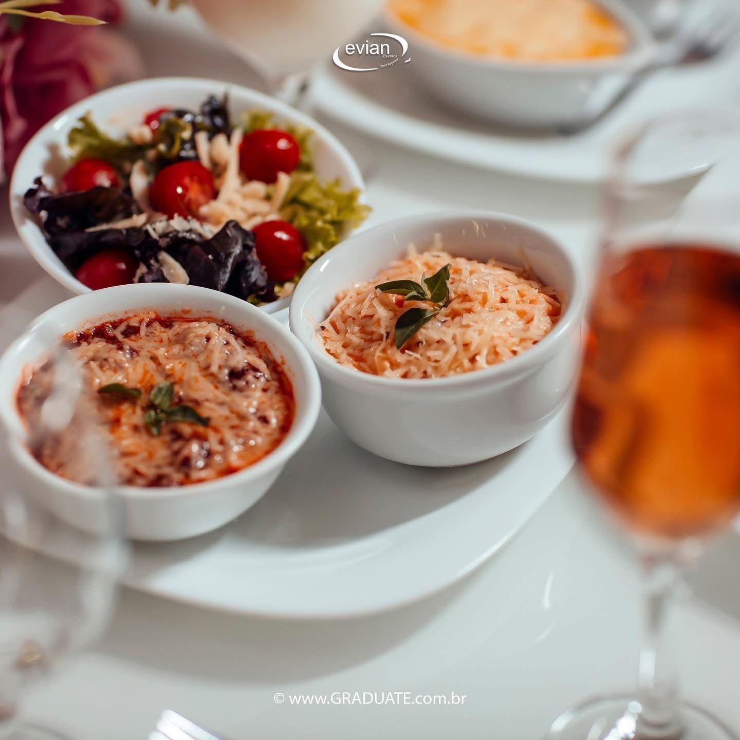 Entrada Evian servida em porções individuais: salada verde com tomates, gratinado de queijo e risoto cremoso ao lado de espumante rosé