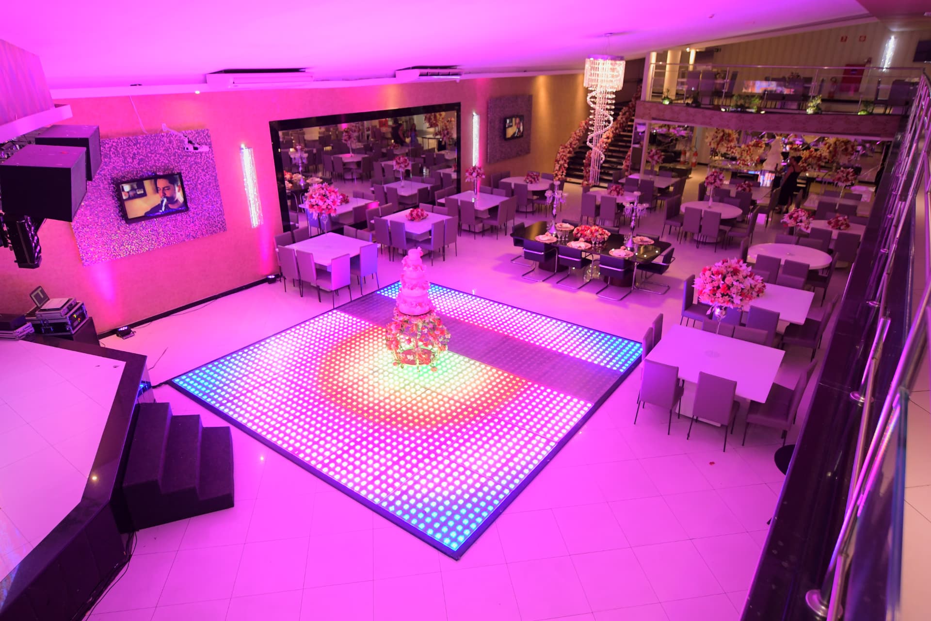 Salão maior do San Pelegrine com iluminação cênica rosa, pista de LED acesa, telão e mesas dos convidados vistas do alto